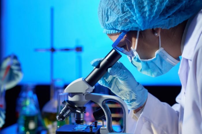 Laboratory scientist in a mask and gloves looking through a microscope